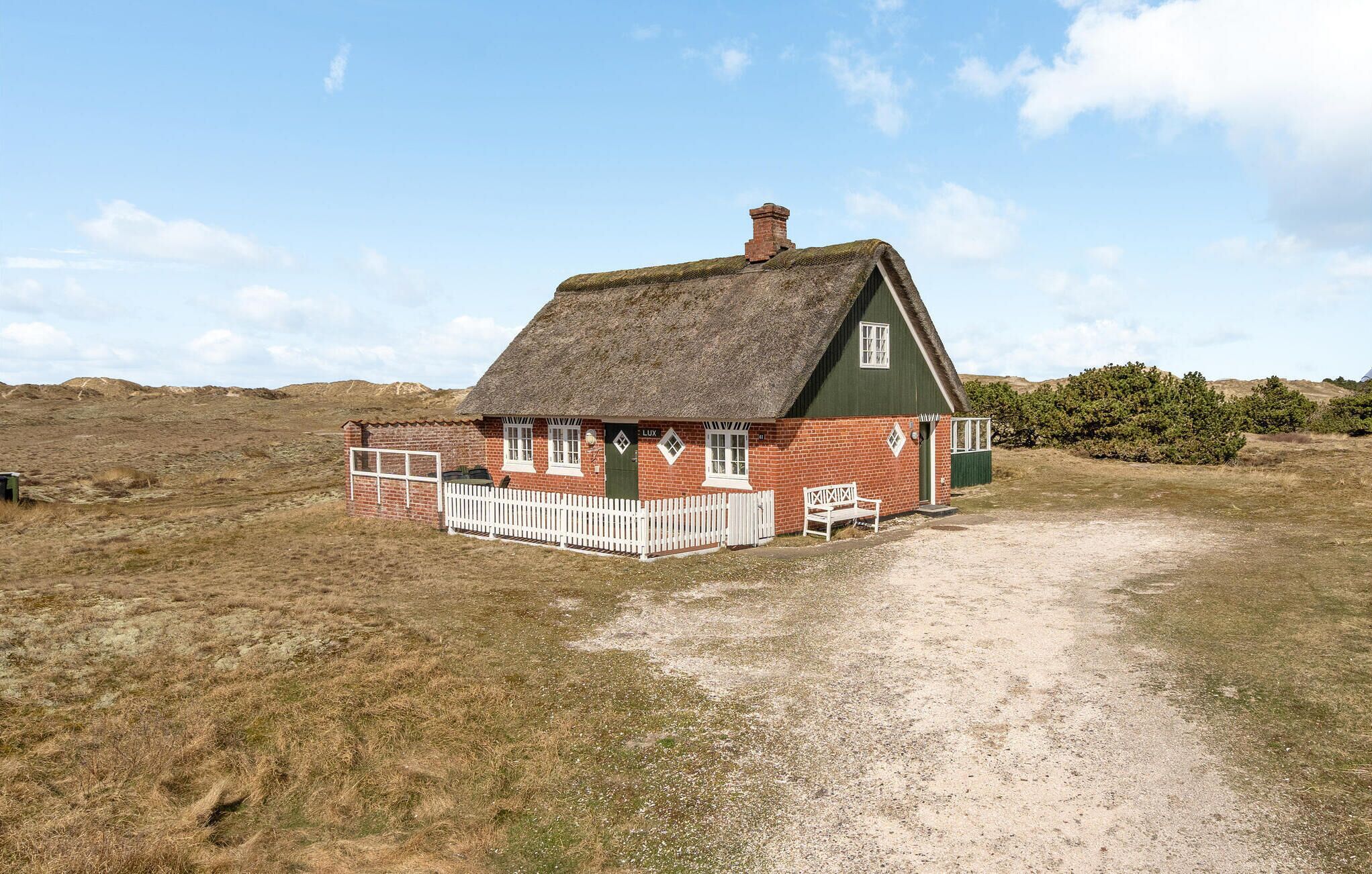 Ein traditionelles Haus mit Strohdach und weißem Zaun, gelegen in ländlicher Umgebung unter einem klaren Himmel.