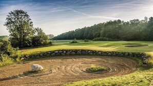 Golfkenttä, jossa on kivimuuri, suuri puu ja kirkas taivas.