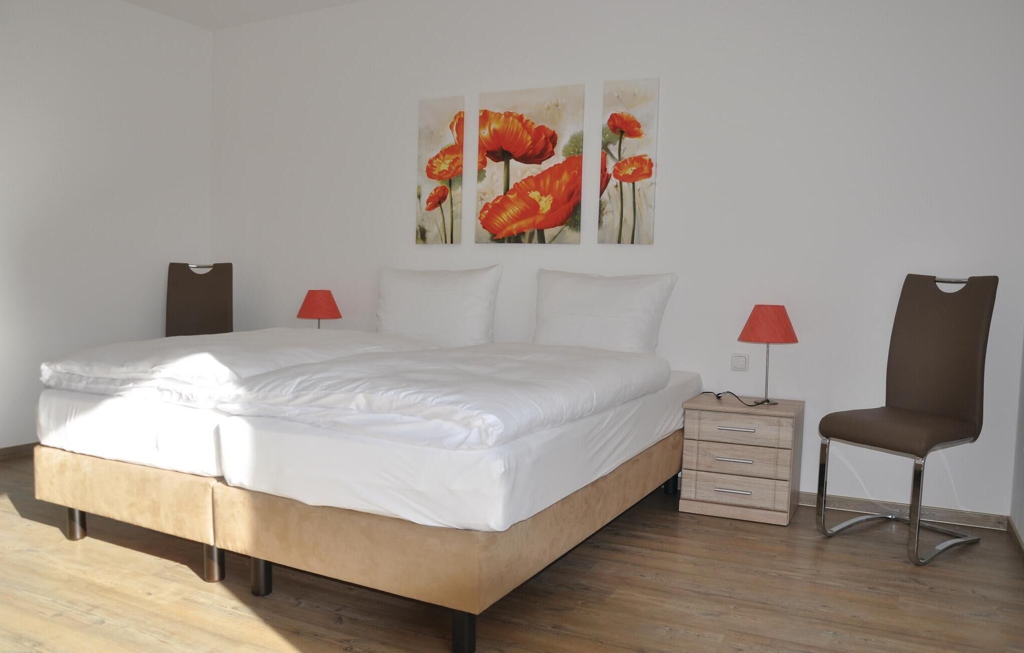 A bedroom with a bed, two bedside tables, a chair, and three wall-mounted paintings.