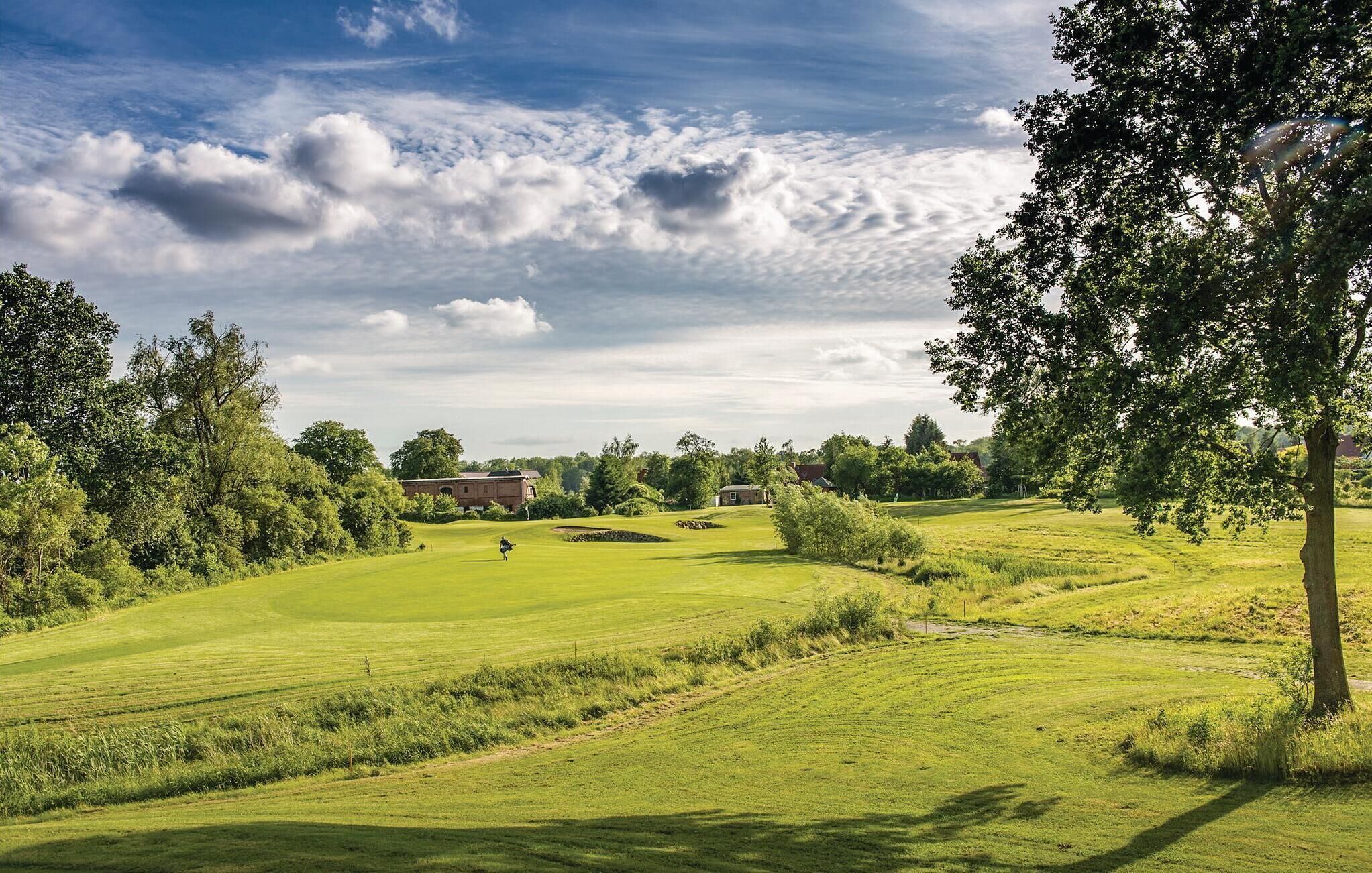 Ein Golfplatz mit sanften, grünen Hügeln, Bäumen und einigen Gebäuden im Hintergrund.