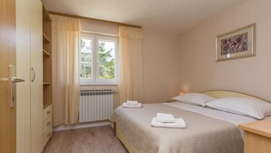 Un dormitorio con cama, un radiador, una ventana con cortinas, una mesita de noche con lámpara y un cuadro en la pared.