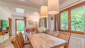 A dining area with a wooden table, chairs, and a vase of flowers. There is a sofa and a television in the background.