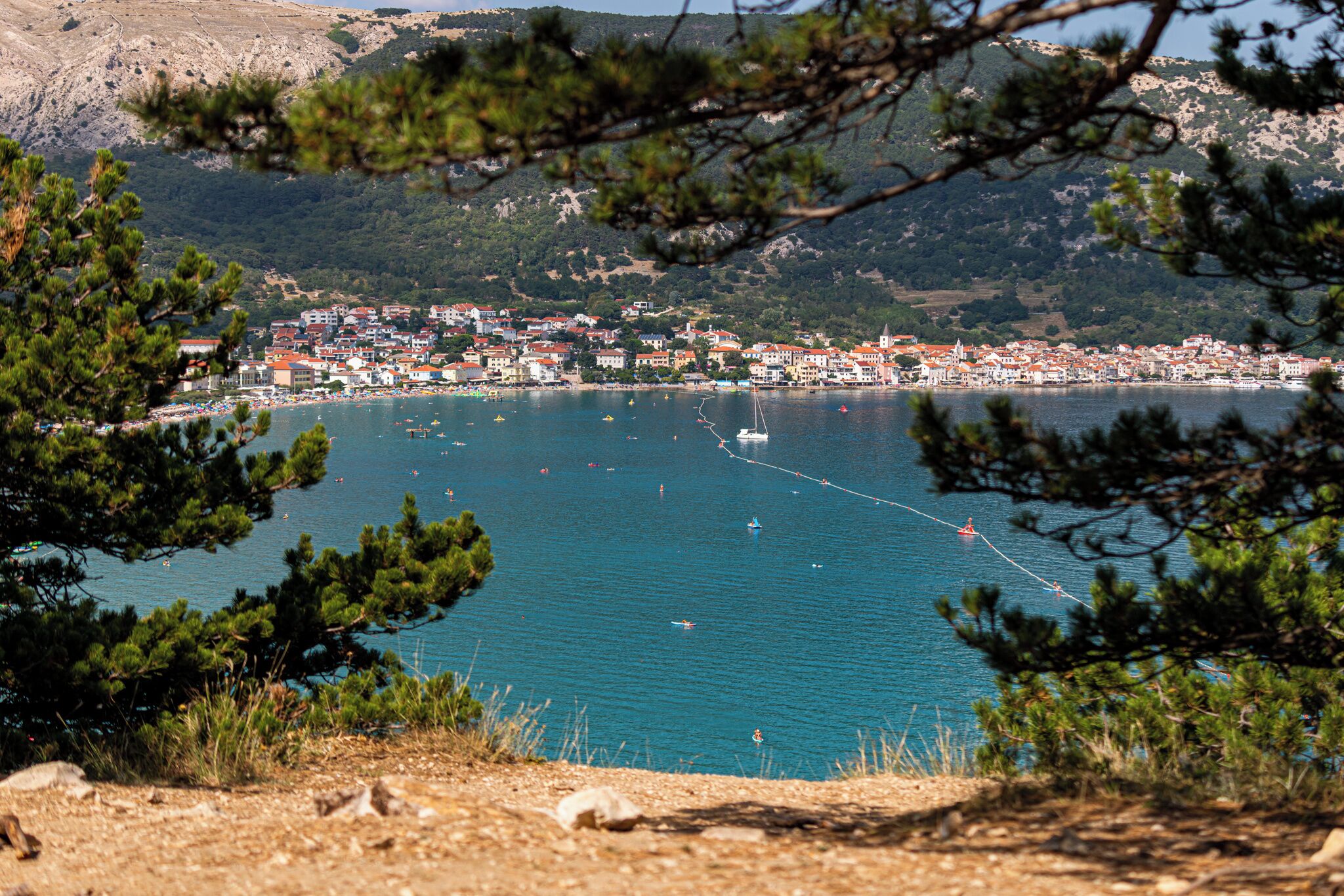 Une ville côtière bordée d’une mer d’un bleu éclatant, entourée de collines et de pins.