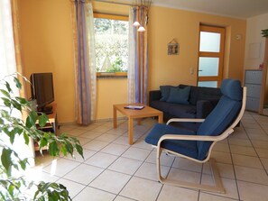 En stue med sofa, stol, lille bord, fjernsyn og et vindue med gardiner.