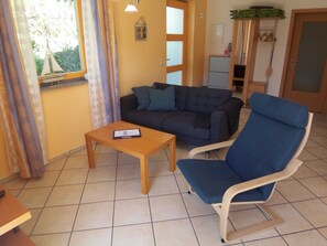 Ein Wohnbereich mit einer Couch, einem blauen Sessel, einem Couchtisch aus Holz und einem Fenster mit Vorhängen.