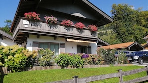 Ein zweistöckiges Gebäude mit einem Balkon, der mit rosa Blüten geschmückt ist, umgeben von gepflegter Bepflanzung und einem Holzzaun.