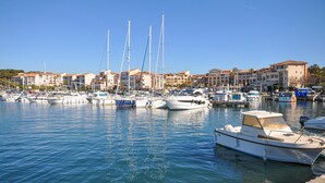 Un port de plaisance où sont amarrés différents types de bateaux, entouré d’immeubles à plusieurs étages dotés de balcons, sous un ciel bleu azur.