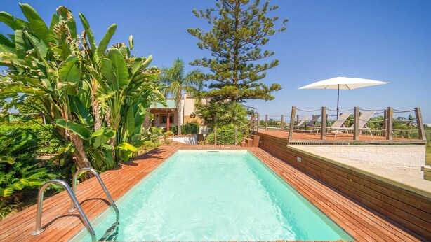 A rectangular swimming pool with a wooden deck surrounding it, a metal ladder, and a white umbrella on the right.