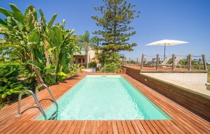 Una piscina rectangular con un muelle de madera alrededor, una escalera metálica y una sombrilla blanca a la derecha.
