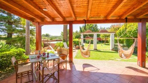 An outdoor patio with a wooden pergola, a dining table with chairs, and two hanging hammocks.