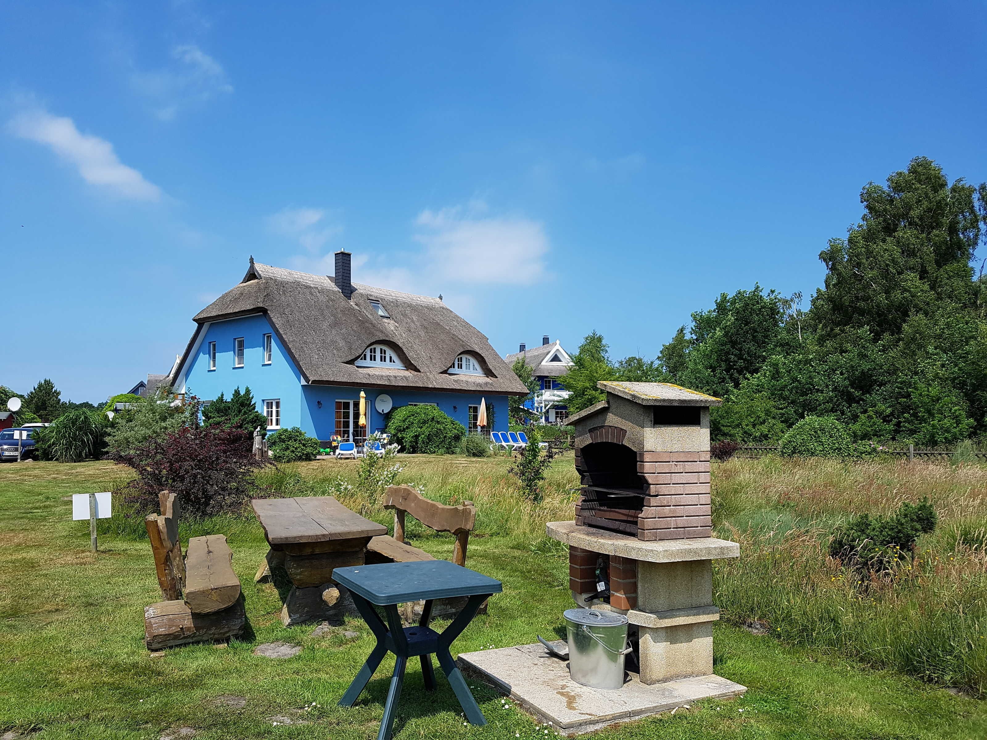 A blue house with a thatched roof, a stone-built outdoor oven, and a picnic table in a grassy area.