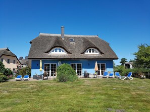 A house with a thatched roof, a blue exterior, and a lawn with lounge chairs.