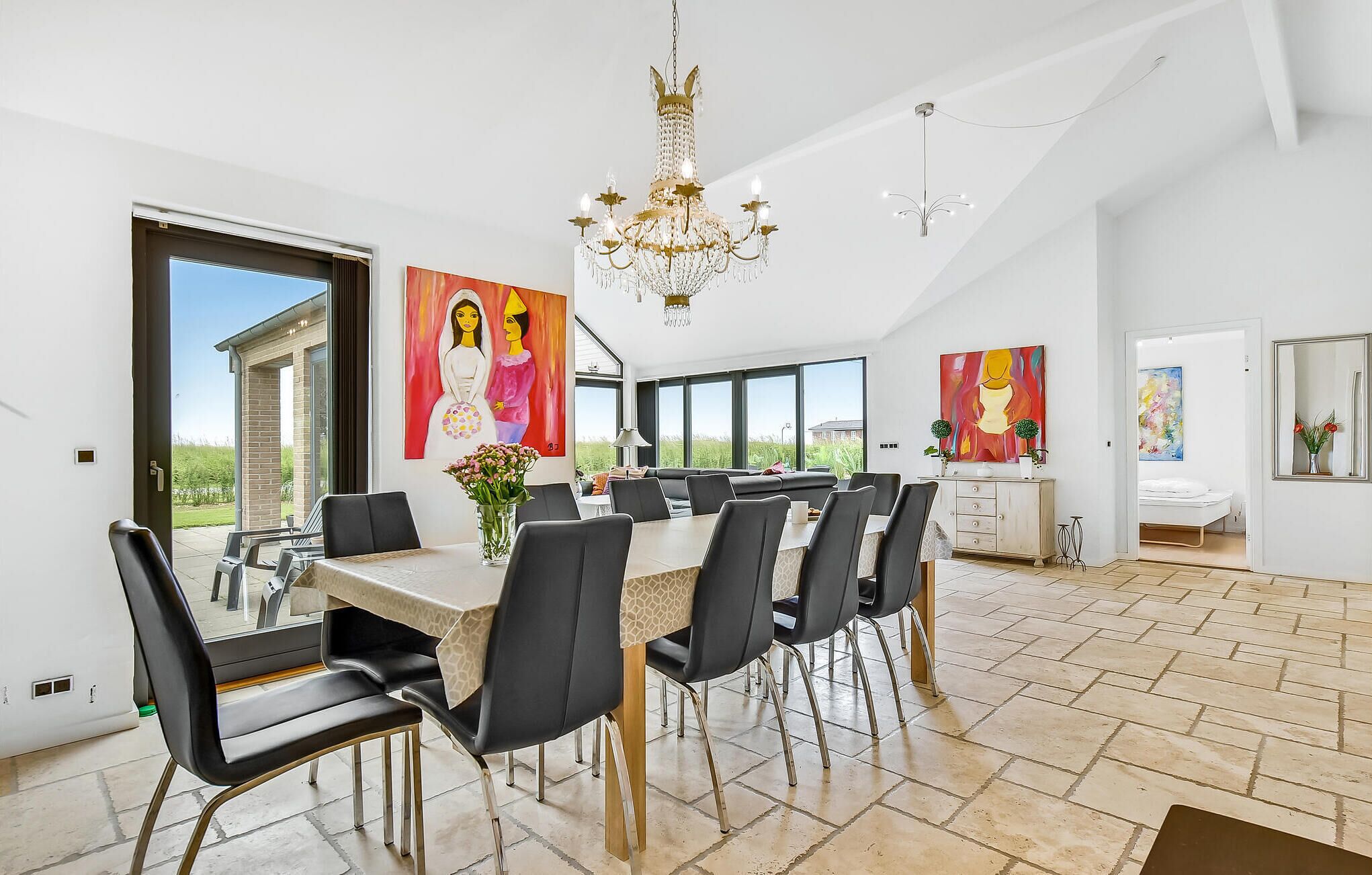 A spacious dining area with a large table, multiple chairs, and a chandelier. There are large windows providing natural light and views of the outdoors.