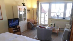 A hotel room with a bed, a television, a sofa, a chair, a table with a bottle and glasses, a cabinet, and a window with curtains. - (129/9) 2-room apartment roof terrace + balcony - apartment house Plückhahn (Kühlungsborn)