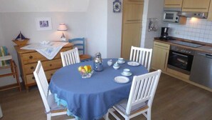 A dining area with a round table covered by a blue tablecloth, surrounded by white chairs. There is a wooden cabinet with a lamp on the table, and a small bowl of fruit on the tablecloth. - (129/9) 2-room apartment roof terrace + balcony - apartment house Plückhahn (Kühlungsborn)