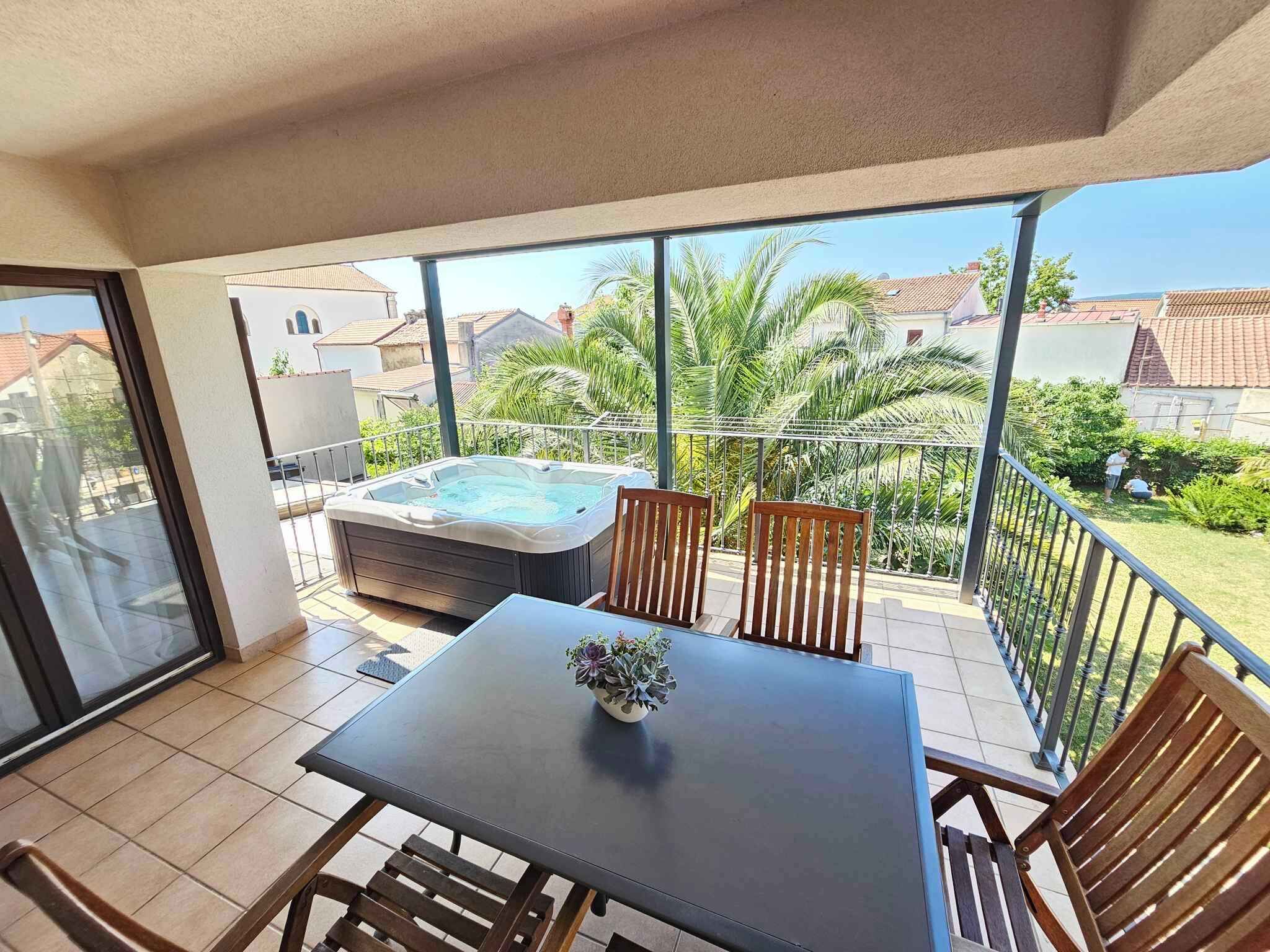 A balcony with a hot tub, a table with chairs, and a view of a residential area with houses and greenery.