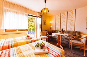 Et værelse med to senge, en sofa, et bord med frugt og udsigt til grønne omgivelser.