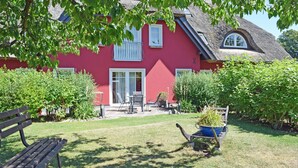 A red house with a thatched roof, a garden with a bench, and a wheelbarrow with plants. - 02 Holiday apartment with terrace - Karola's country house under the thatched roof (Mönchgut)