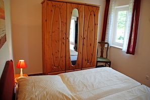 A bedroom with a bed, a wooden wardrobe, a chair, a lamp, and a window. - 02 Holiday apartment with terrace - Karola's country house under the thatched roof (Mönchgut)