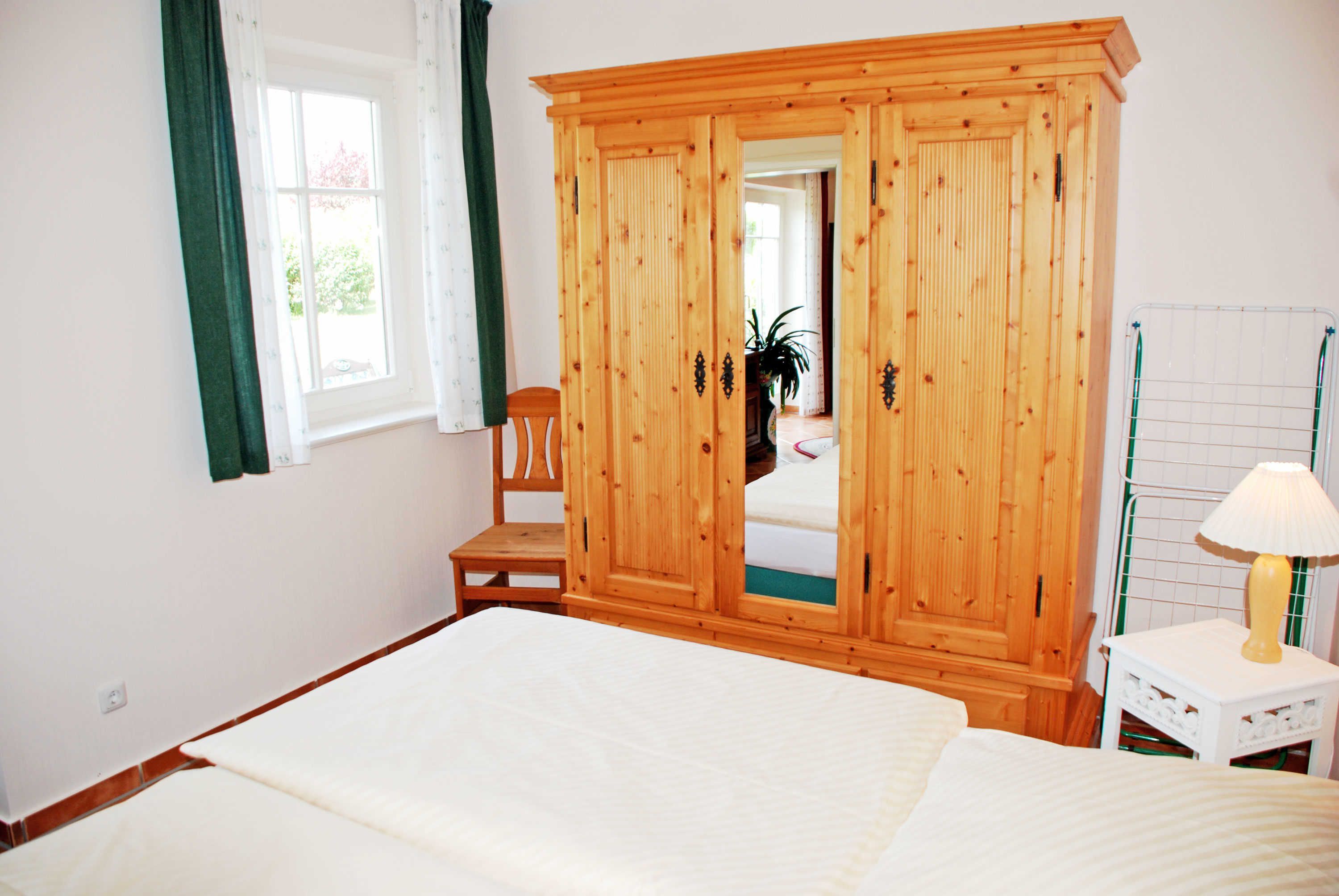 A bedroom with a wooden wardrobe, a bed, a chair, and a small table with a lamp.