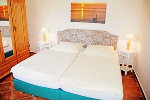 A double bed with a patterned headboard, two bedside tables with lamps, a wooden wardrobe, and a sunset painting on the wall.