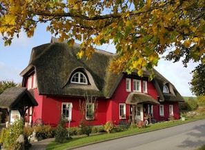 A red house with a thatched roof, surrounded by greenery and a clear sky. - 02 Holiday apartment with terrace - Karola's country house under the thatched roof (Mönchgut)