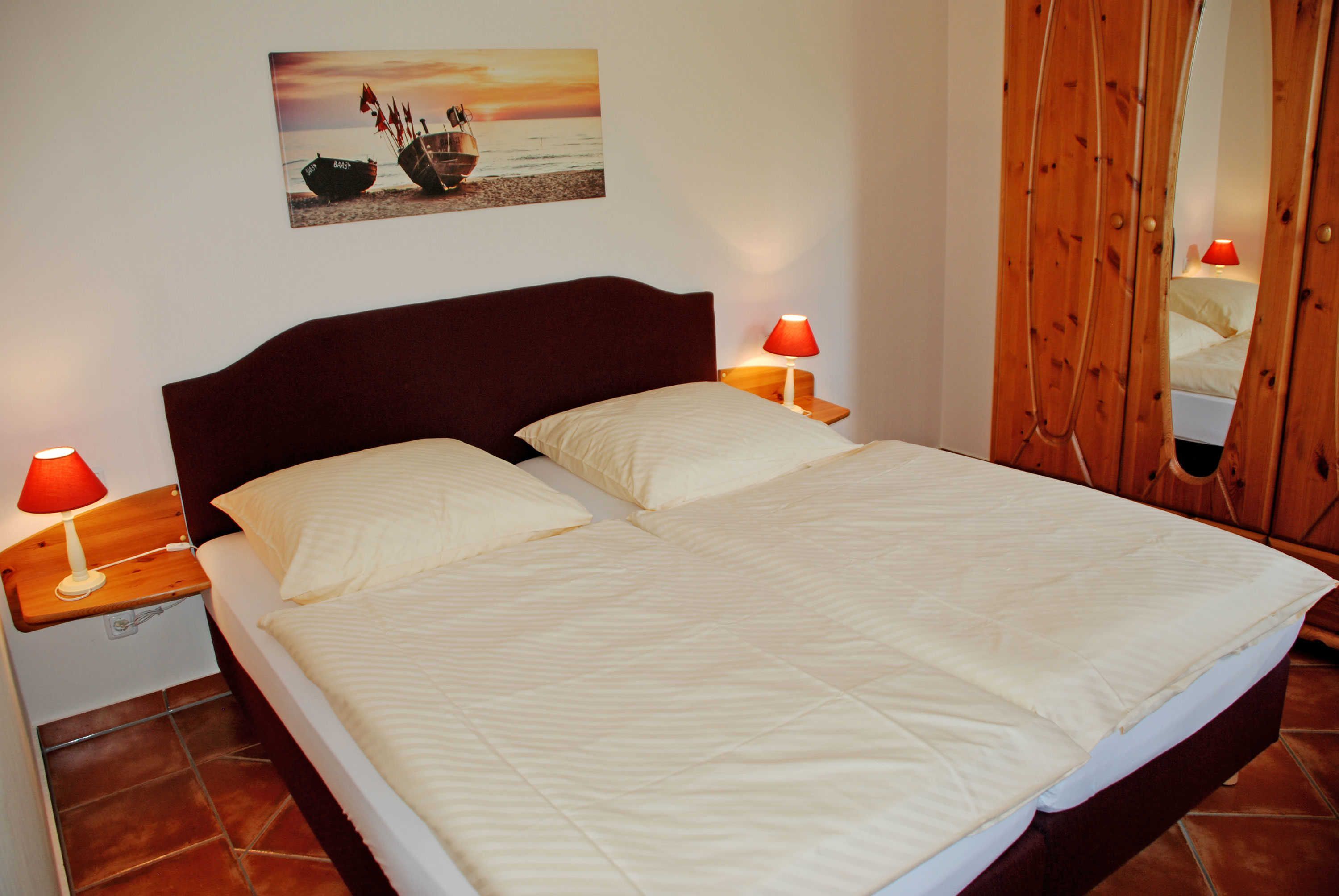 Ein Bett mit zwei Kissen, ein Nachttisch mit roter Lampe, ein Holz-Kopfteil und ein an der Wand hängendes Bild von Booten.