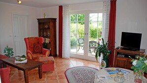 A living room with a sofa, armchair, wooden cabinet, television, and a table with a book and a vase. - 02 Holiday apartment with terrace - Karola's country house under the thatched roof (Mönchgut)
