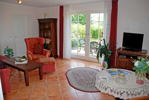 A living room with a sofa, armchair, wooden cabinet, television, and a table with a book and a vase. - 02 Holiday apartment with terrace - Karola's country house under the thatched roof (Mönchgut)