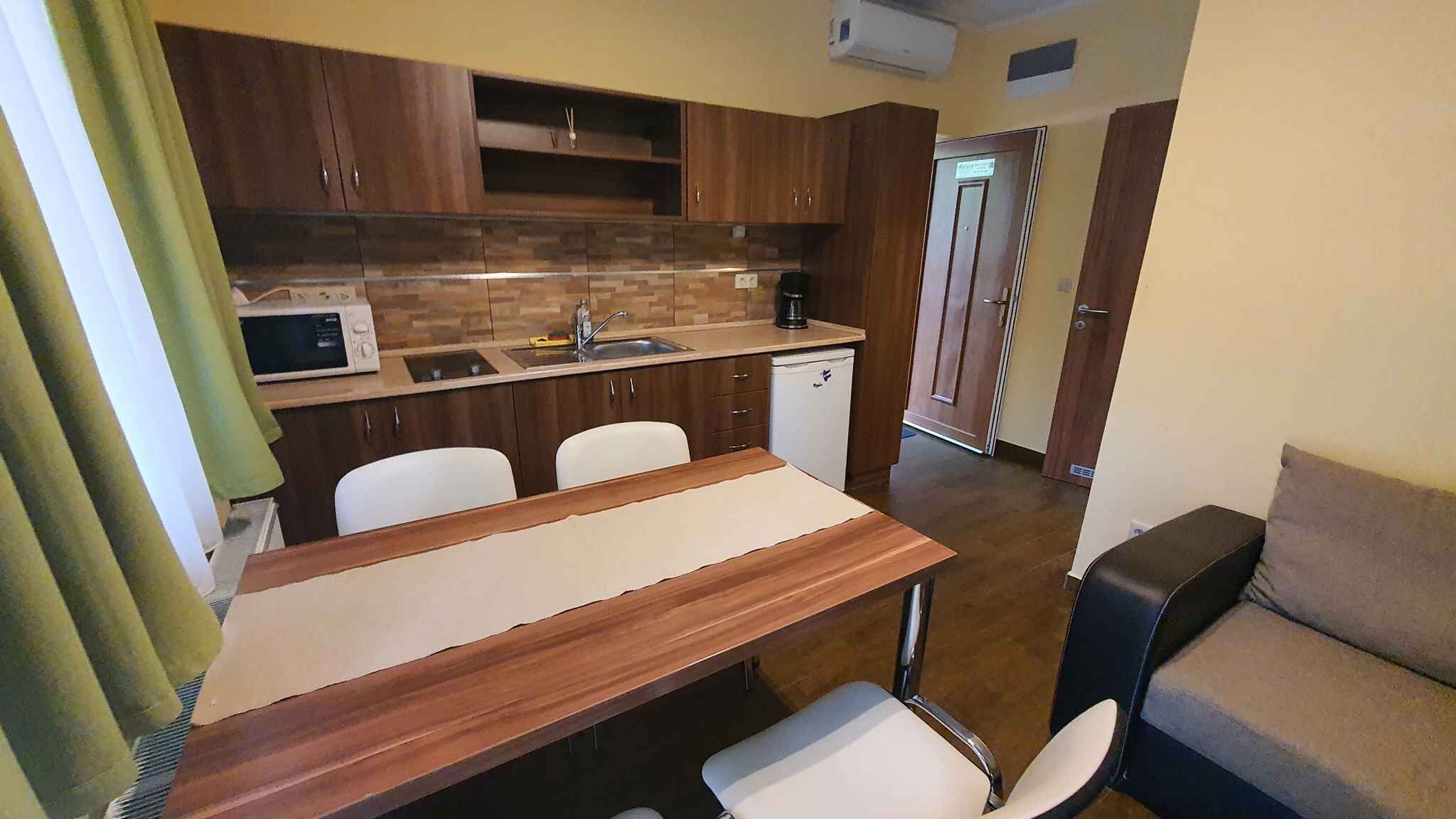 A compact kitchen with a microwave, sink, and white cabinets. There is a wooden table and two chairs. A small refrigerator is visible in the background.
