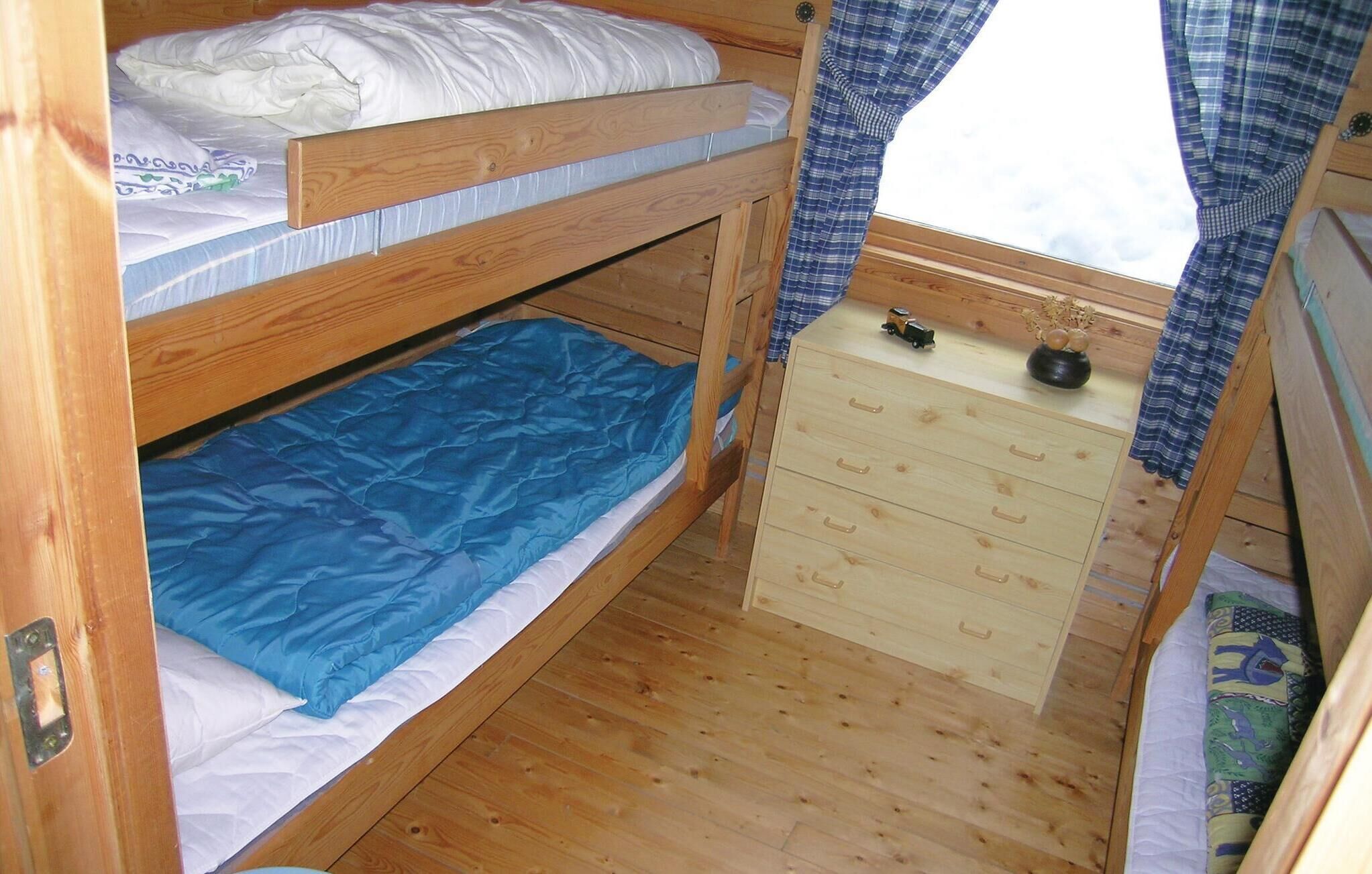 A bunk bed room with a wooden floor, a small wooden dresser with drawers, and a window with curtains.