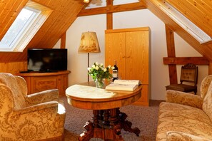 Ein gemütliches Zimmer mit Holzbalkendecke, einem Fernseher, einem runden Holztisch mit Blumenvase und zwei gemusterten Sesseln.