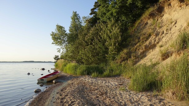 A sandy beach with a red boat and a rocky shoreline. - Small cottage on the forest and hunting lodge - Forest & Jagdgut (Lütow)