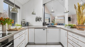 A modern kitchen with white cabinetry, a built-in dishwasher, and a large island with a sink. There are hanging knives, a coffee maker, and a potted plant on the counter. - Gorgeous home in Tisvildeleje with sauna (Tisvildeleje)