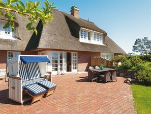 A thatched-roof house with a brick patio, a lounge chair, and a table with chairs.