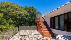 A tiled patio with a staircase leading to a wooden deck, surrounded by a metal fence and lush greenery.