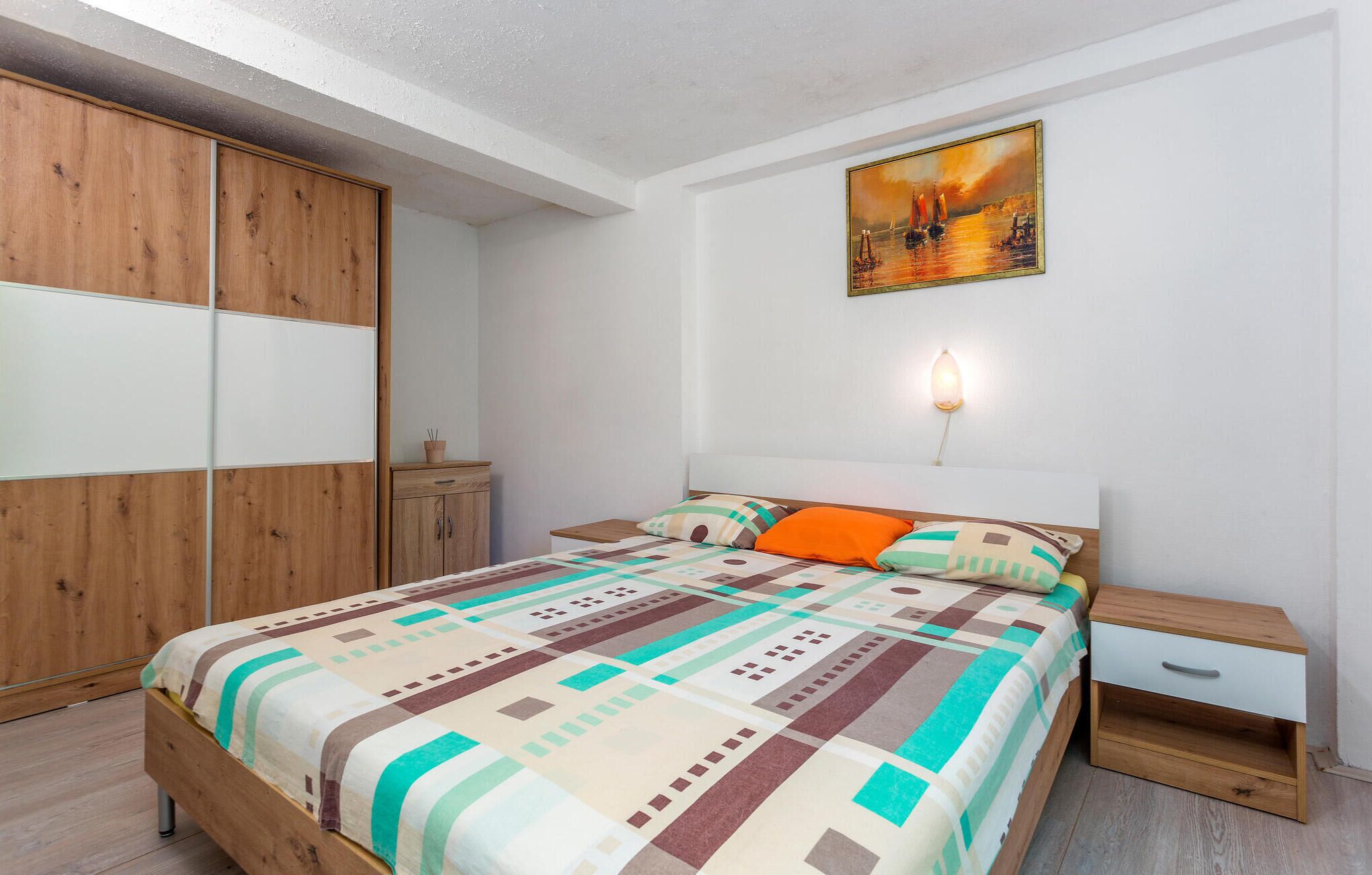 Een slaapkamer met een bed, nachtkastje, houten garderobe, schilderij en een wandlamp.