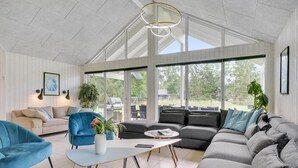 A spacious living room with a large sectional sofa, a blue armchair, a coffee table, and a dining table. There are large windows providing a view of the outdoors. - Gorgeous home in Frederiksværk with sauna (Frederiksværk)