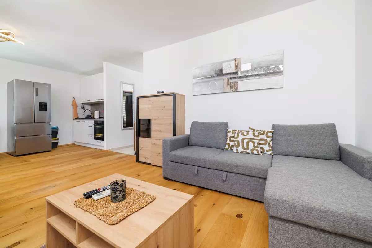 A modern living room with a grey sofa, wooden coffee table, and a kitchen area with a fridge and cabinets.