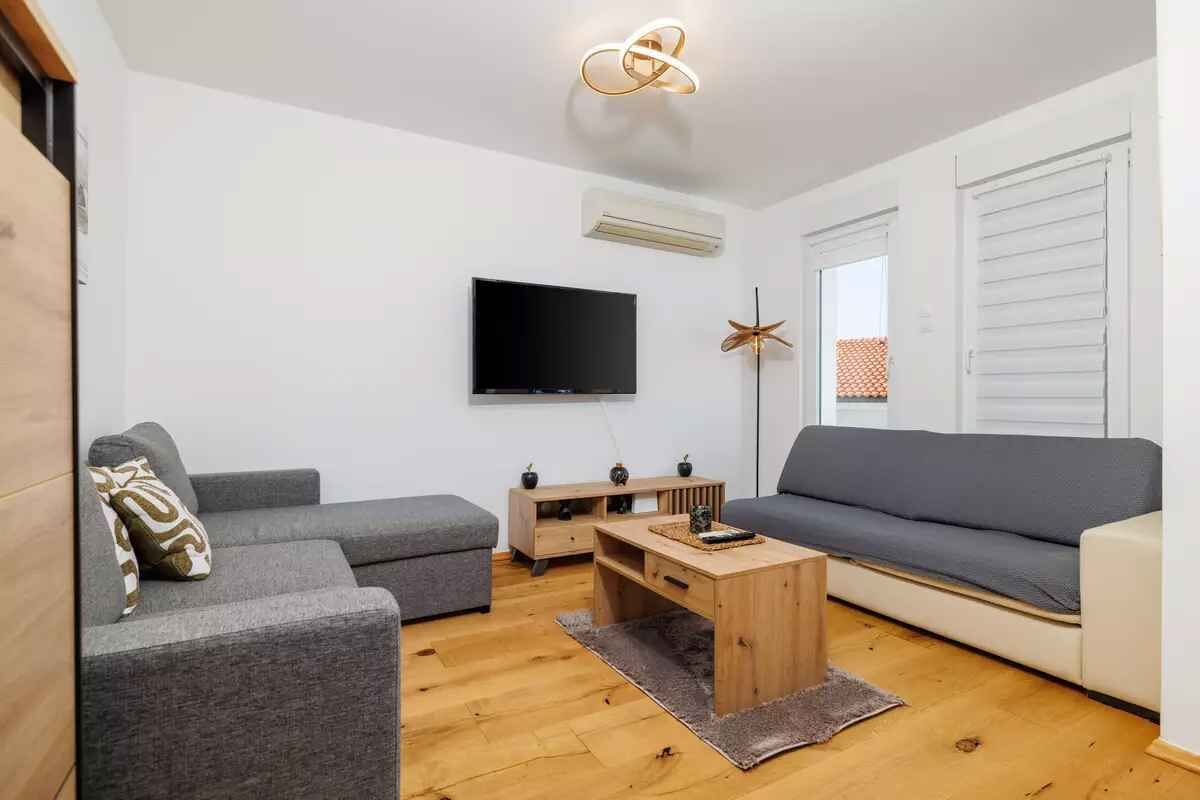 A modern living room with a flat-screen TV, grey sofas, a wooden coffee table, and a ceiling light fixture.