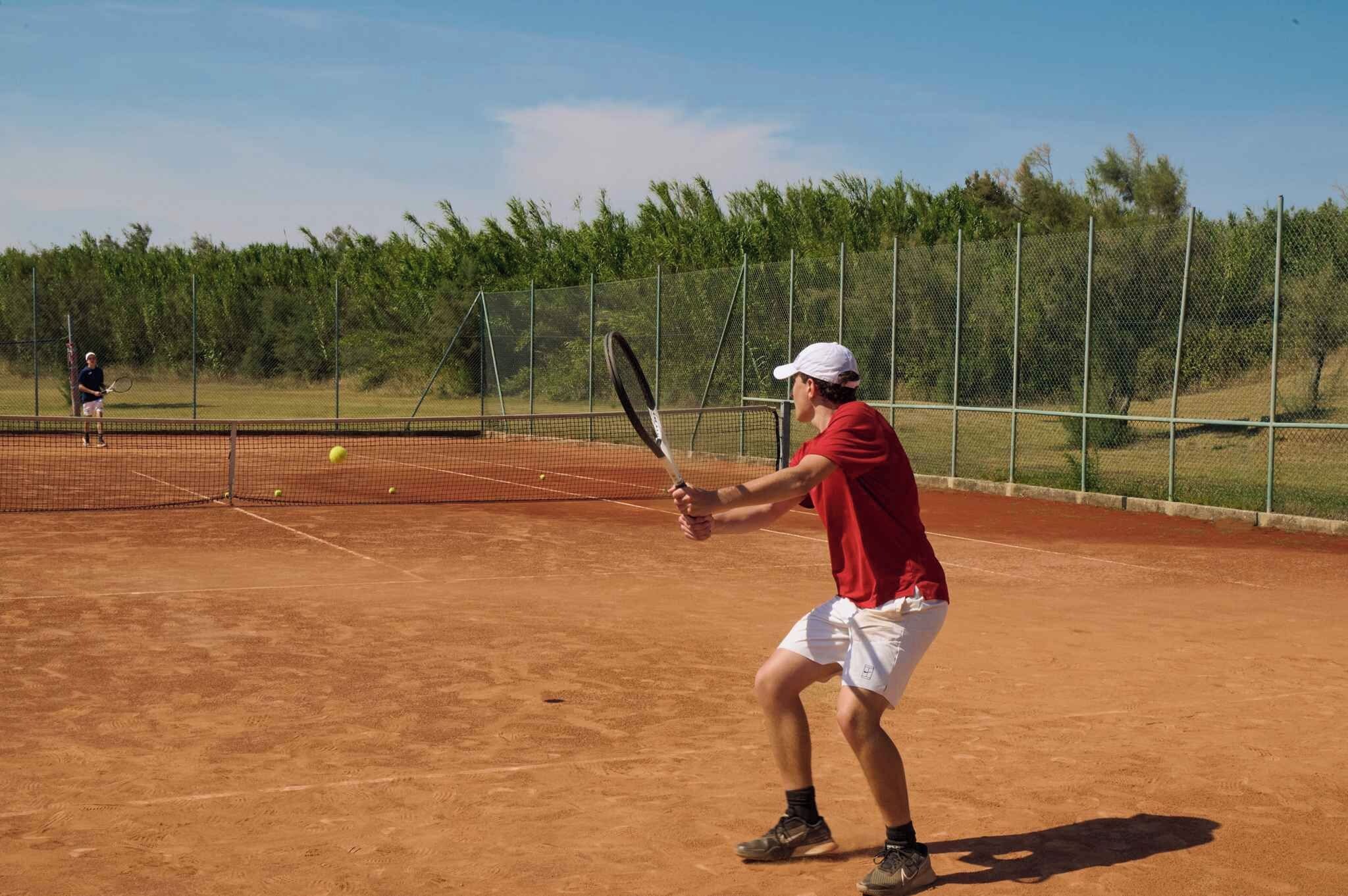 Una cancha de tenis con un jugador con camisa roja y pantalon corto blanco, una red y una valla al fondo.