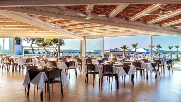 Un amplio comedor interior con mesas y sillas, grandes ventanales que ofrecen vista a una playa y palmeras.