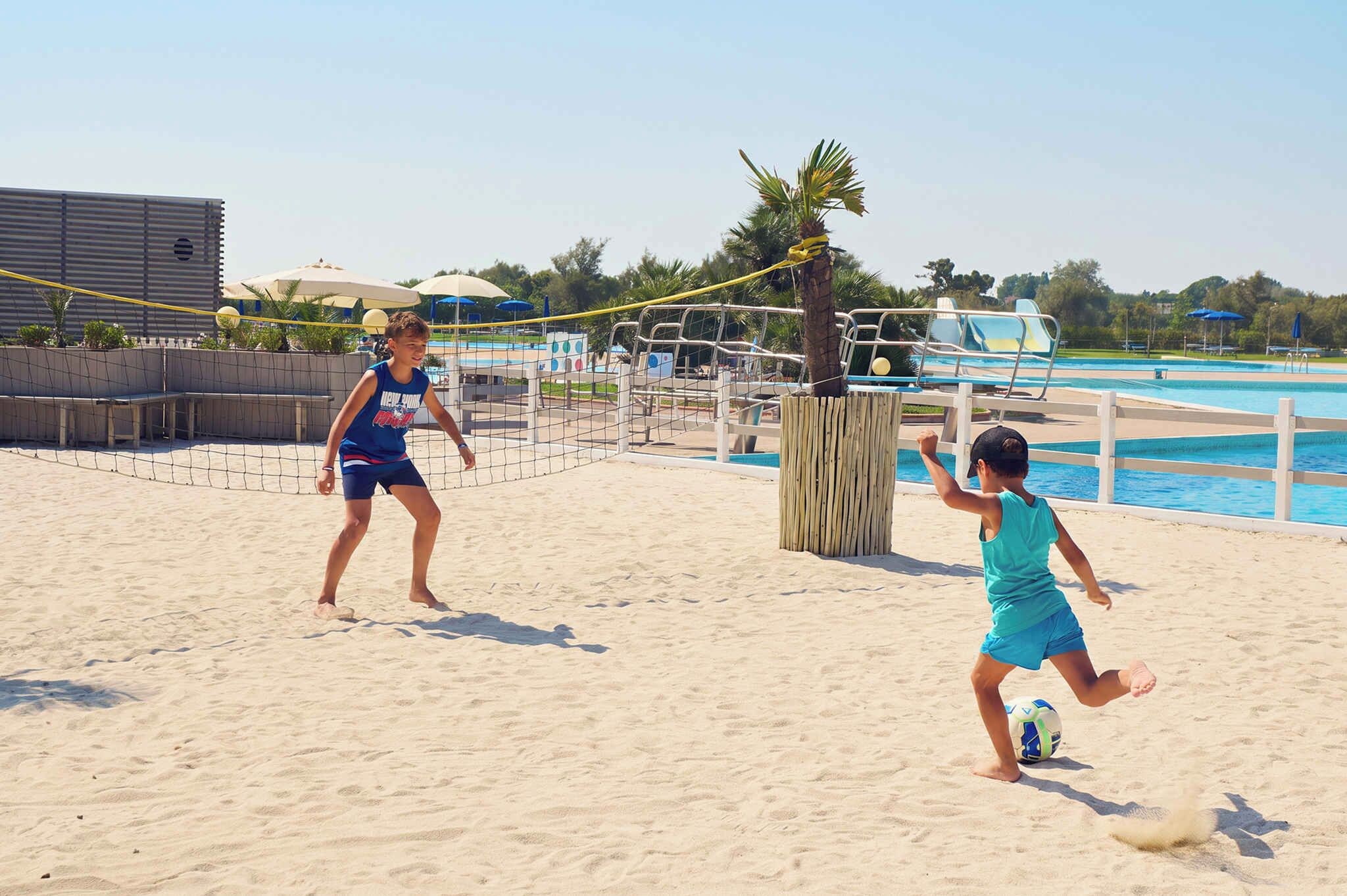 Two children playing beach volleyball on a sandy court with a pool and umbrellas in the background.