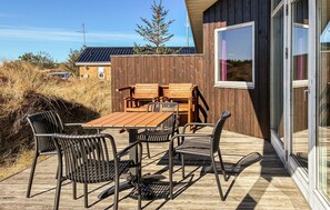 En udendørs terrasse med et træbord og stole, en træterrasse samt en bygning med store vinduer.