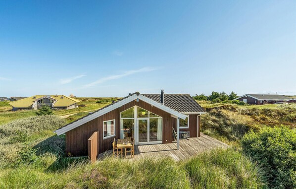 Et træhus med skråt tag og store glasdøre, omgivet af højt græs og andre huse i baggrunden.