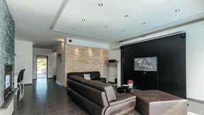 Un salon moderne avec un canapé en cuir, un pouf noir, un mur en pierre, des spots encastrés au plafond et un téléviseur à écran plat fixé au mur.