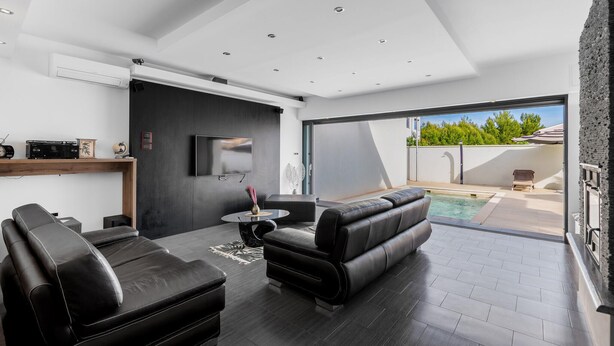 Un salon moderne avec un canapé en cuir noir, une petite table ronde et un téléviseur à écran plat fixé au mur. Une grande porte-fenêtre coulissante donne accès à un espace extérieur avec une piscine.
