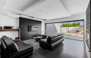 A modern living room with a black leather sofa, a small round table, and a flat-screen TV mounted on the wall. There is a large sliding glass door leading to an outdoor area with a pool. - Gorgeous home in Vrsi Mulo with private swimming pool, can be inside or outside (Vrsi Mulo)