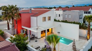 Une maison moderne à deux étages, dotée d’une piscine, d’une façade rouge et blanche, et offrant une vue sur les montagnes en arrière-plan.