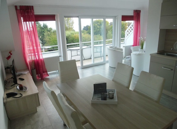Un espace repas moderne avec une table dressée pour deux personnes, un coin cuisine équipé d’un évier et de placards, et un balcon avec une table et des chaises.